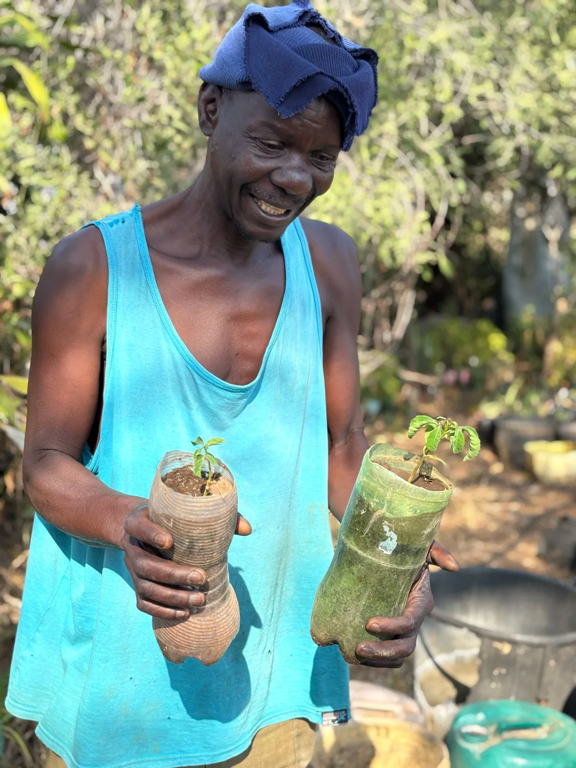 Charles Mdluli at Local Plants nursery, smiling, holding two young saplings in upcycled-bottle planters.