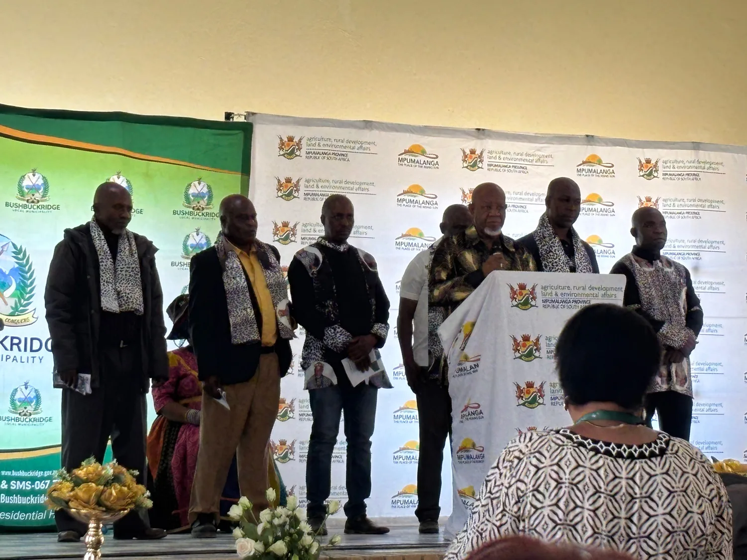 Kgoshi L Mokoena, Chairperson of the Mpumalanga House of Traditional Leaders and Khoisan Leaders, addresses Provincial Arbor Day 2025 at Oakley Community Hall, Bushbuckridge. Fellow traditional leaders flank him on stage; DARDLEA (Mpumalanga Agriculture, Rural Development, Land & Environmental Affairs) and Bushbuckridge Local Municipality branding frame the podium.