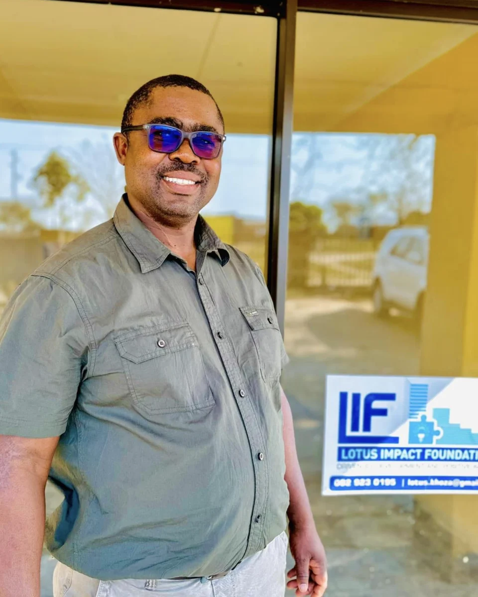 Lotus Khoza standing in front of the Lotus Impact Foundation office, wearing an olive-grey shirt and blue-tinted sunglasses, smiling warmly. The Lotus Impact Foundation (LIF) signage is visible on the window behind him.