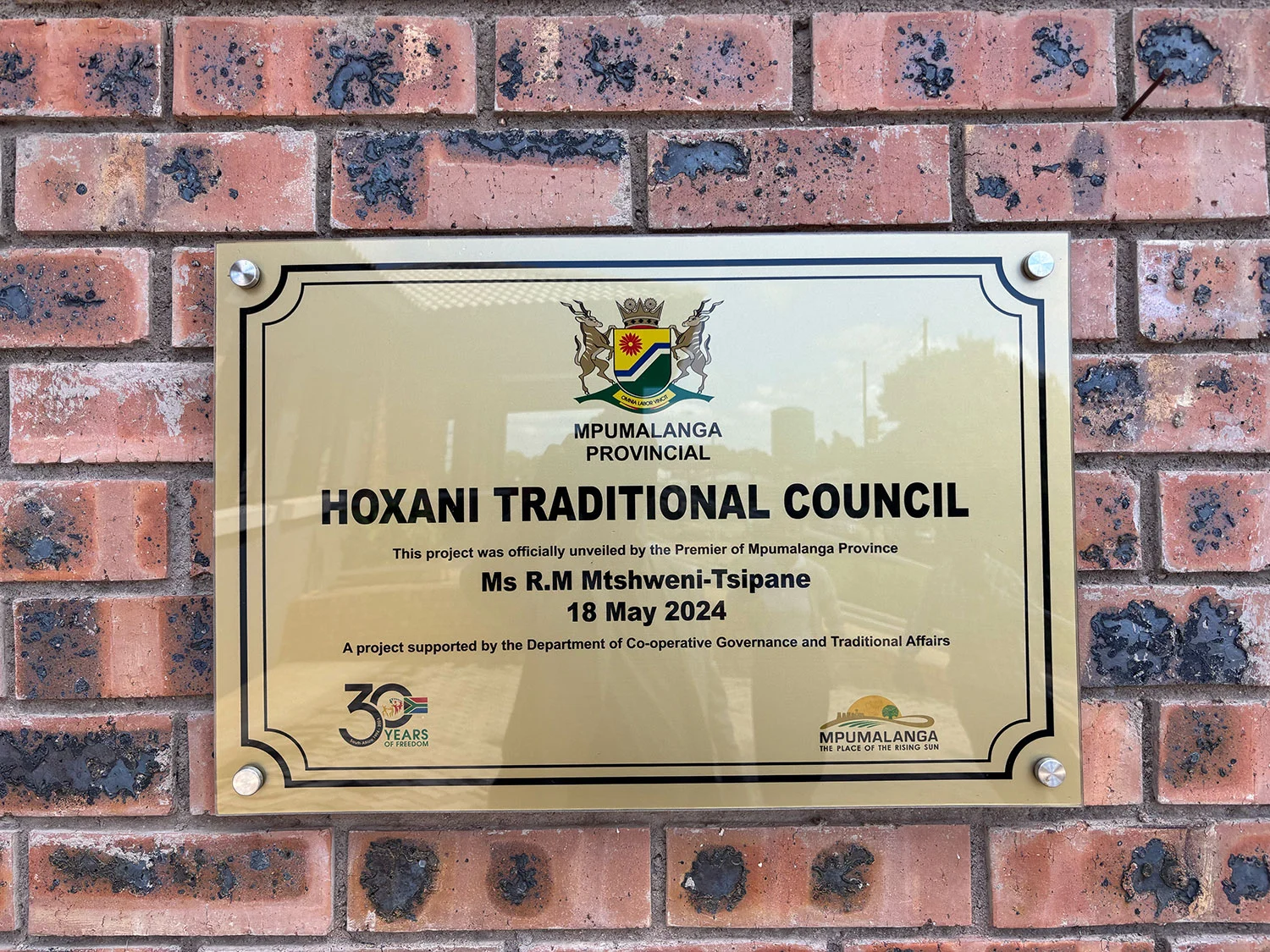 Brass commemorative plaque mounted on a brick wall reading 'MPUMALANGA PROVINCIAL · HOXANI TRADITIONAL COUNCIL · This project was officially unveiled by the Premier of Mpumalanga Province Ms R.M Mtshweni-Tsipane · 18 May 2024 · A project supported by the Department of Co-operative Governance and Traditional Affairs'. Mpumalanga provincial crest and 30 Years of Freedom badge visible on the plaque.