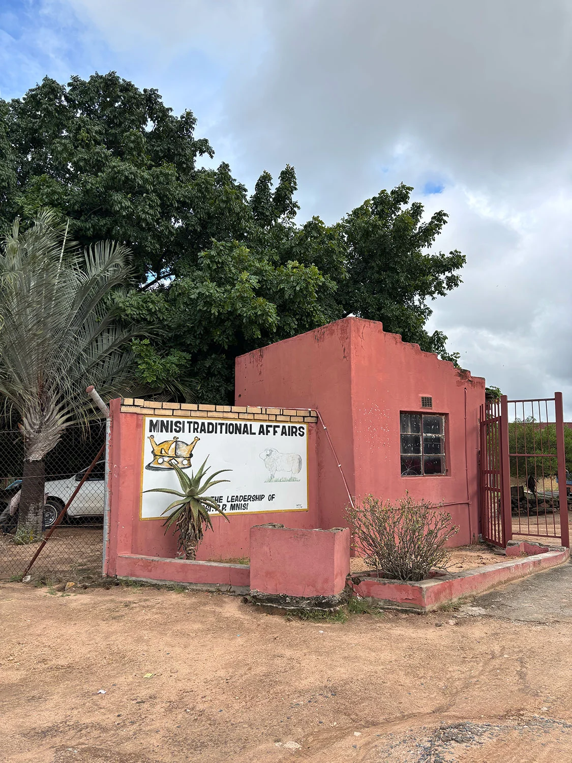 The Mnisi Traditional Affairs building — a pink-painted brick structure with a painted sign reading 'MNISI TRADITIONAL AFFAIRS — UNDER THE LEADERSHIP OF INKOSI P. MNISI' and a crown emblem. A desert-rose palm and aloe grow in front; a fence runs along the boundary.