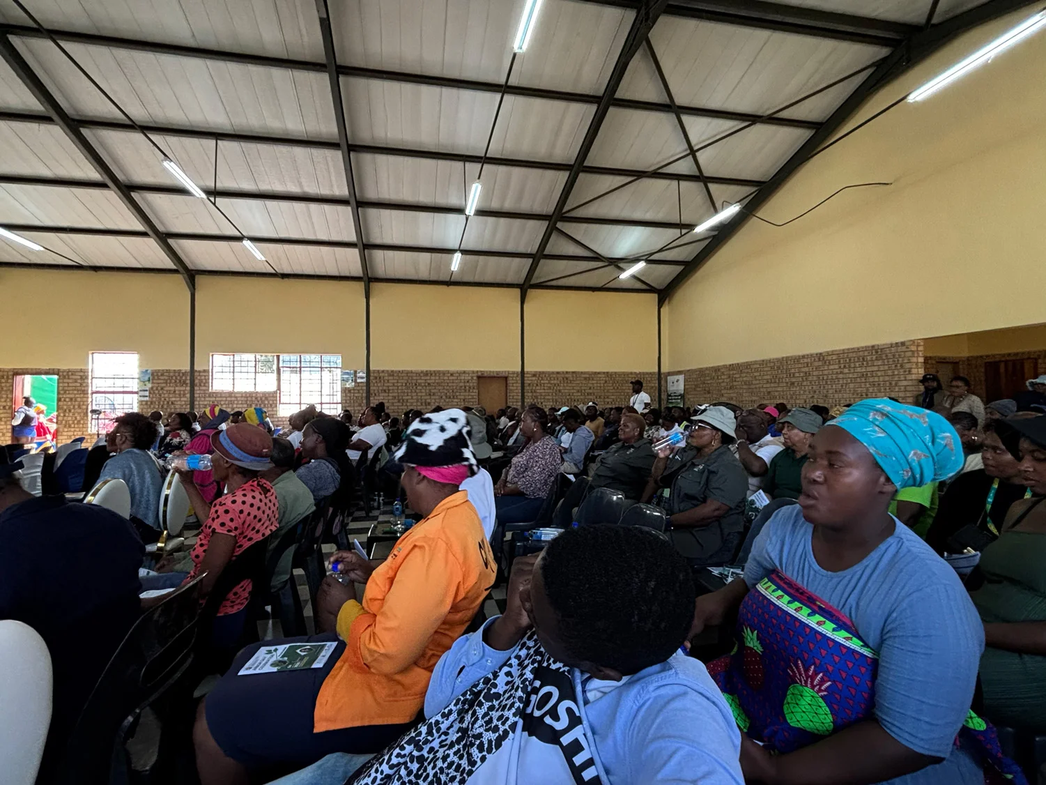 Oakley Community Hall filled with community members — women in patterned dress and headscarves, elders, ward representatives and farmers — seated in rows, attending the Provincial Arbor Day 2025 programme at which Kgoshi L Mokoena and Save the Sand both addressed. Iron-truss roof and yellow-washed brick walls.