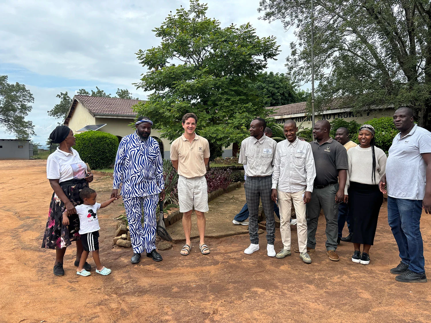 Group photograph at a Free Prior Informed Consent (FPIC) meeting with the Jongilanga Traditional Authority. The Inkosi wears zebra-pattern ceremonial attire and stands beside Save the Sand CEO Richie Laburn, flanked by councillors, a woman in patterned Shangaan dress holding a child, and community members. A traditional council building with a pitched roof and red door sits behind; red-earth yard underfoot.