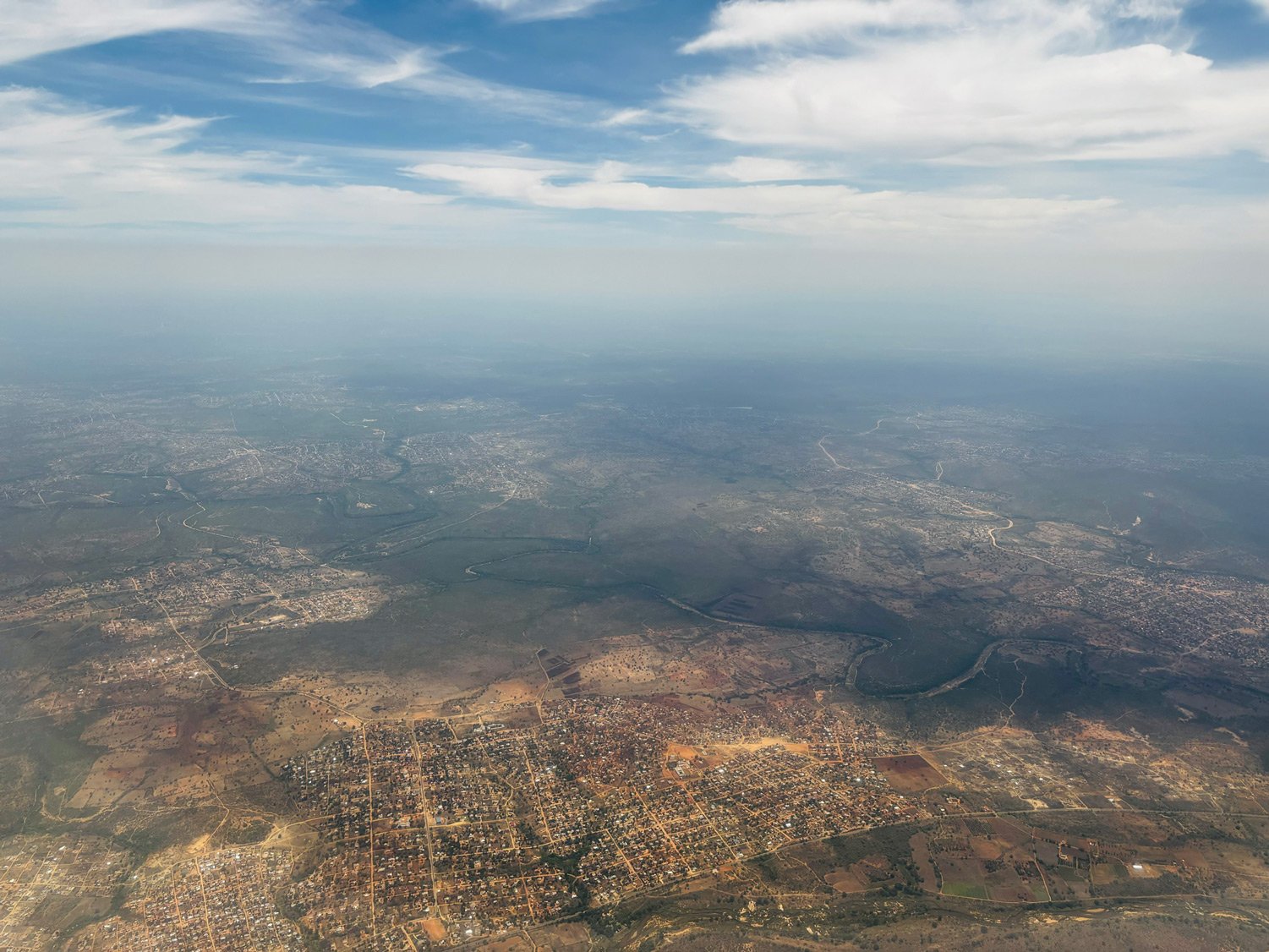 High aerial of the Sabie-Sand catchment — rural settlements spreading across the landscape toward distant ridges and the Drakensberg escarpment, hazy horizon line meeting sky.